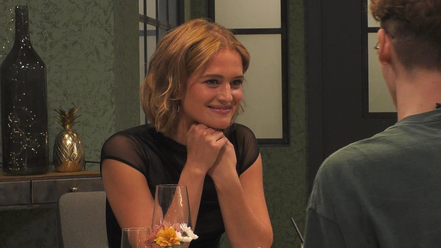 Promi First Dates 2025: Ex-Bachelor Paul Janke, Xenia von Sachsen und weitere Promis auf Liebessuche 3 Chiara Moon Horst, eine junge Frau mit kinnlangem, welligem blondem Haar, sitzt an einem Tisch in einem stilvollen Restaurant. Sie trägt ein schwarzes, leicht durchsichtiges Oberteil und lächelt freundlich, während sie ihre Hände unter ihr Kinn stützt. Im Vordergrund ist die Silhouette ihres Gesprächspartners zu sehen, im Hintergrund eine goldene Ananas-Dekoration und stilvolle Beleuchtung.