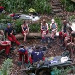 Eine Gruppe von Dschungelcamp-Kandidaten sitzt in einem Kreis auf Baumstämmen im australischen Dschungel. Sie tragen typische rote Hosen und blaue Shirts mit ihren Namen. Rund um das Camp sind Schlafplätze und Ausrüstungsgegenstände zu sehen. Die Atmosphäre wirkt gespannt, während die Kandidaten diskutieren.
