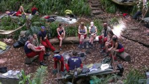 Eine Gruppe von Dschungelcamp-Kandidaten sitzt in einem Kreis auf Baumstämmen im australischen Dschungel. Sie tragen typische rote Hosen und blaue Shirts mit ihren Namen. Rund um das Camp sind Schlafplätze und Ausrüstungsgegenstände zu sehen. Die Atmosphäre wirkt gespannt, während die Kandidaten diskutieren.