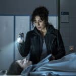 Harry Möller (gespielt von Maria Ketikidou) steht mit einer Taschenlampe in der Hand vor der zugedeckten Leiche von Mirja Grabowski (Franziska von Harsdorf). Der ernste Ausdruck in ihrem Gesicht spiegelt ihre Fassungslosigkeit wider. Die Szene spielt in einem düsteren Raum, vermutlich in der Pathologie, und deutet auf die Dramatik der Handlung im Film „Großstadtrevier – Im Moment der Angst“ hin.