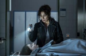 Harry Möller (gespielt von Maria Ketikidou) steht mit einer Taschenlampe in der Hand vor der zugedeckten Leiche von Mirja Grabowski (Franziska von Harsdorf). Der ernste Ausdruck in ihrem Gesicht spiegelt ihre Fassungslosigkeit wider. Die Szene spielt in einem düsteren Raum, vermutlich in der Pathologie, und deutet auf die Dramatik der Handlung im Film „Großstadtrevier – Im Moment der Angst“ hin.