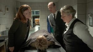 Die Bestatter Frau Bleckmann (Regine Hentschel) und Herr Töteberg (Stephan A. Tölle) stehen in einem kalten Raum neben Friseurin Grit Hartmann (Julika Jenkins), während sie gemeinsam eine zugedeckte Leiche auf einer Bahre betrachten.