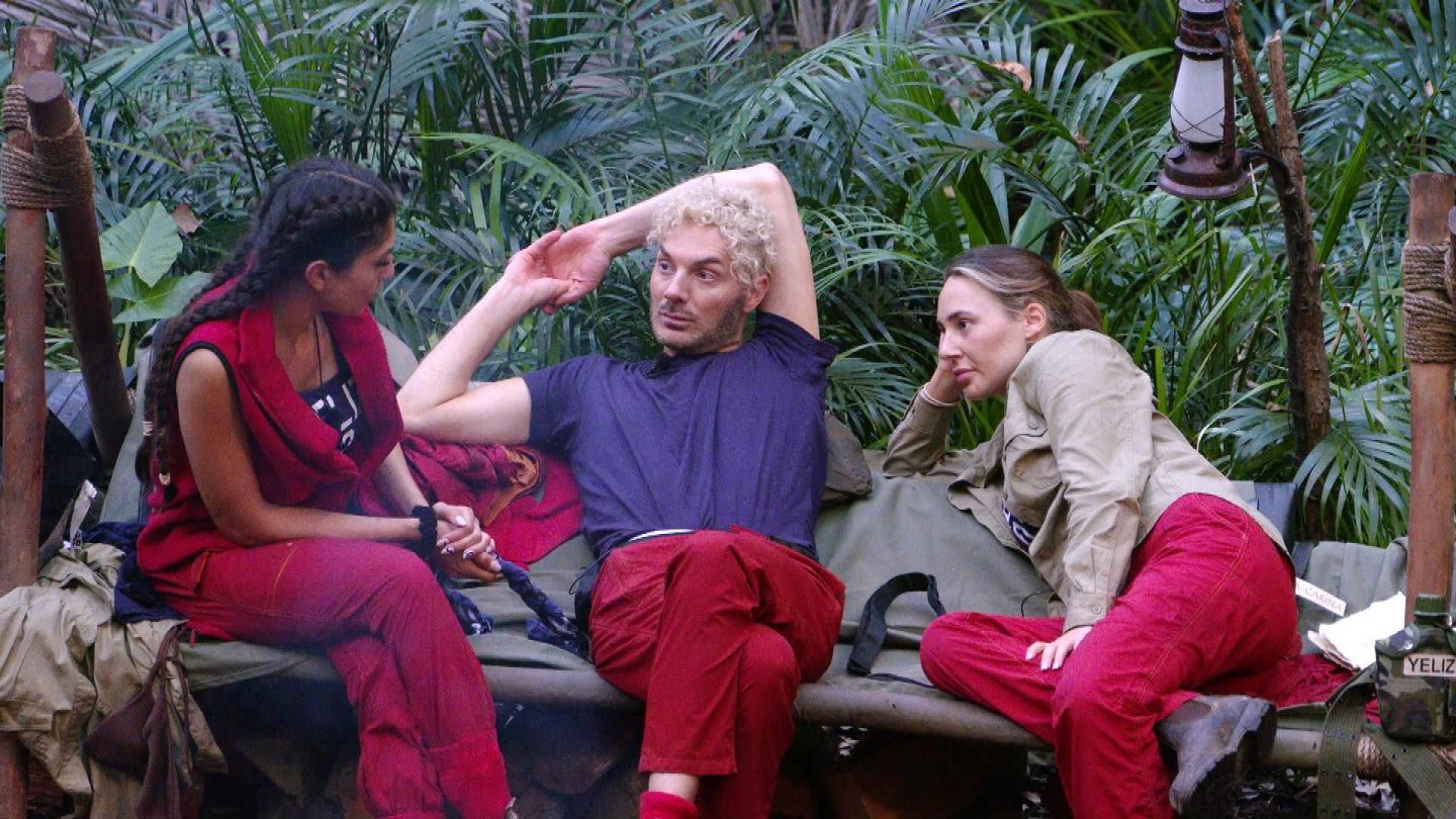 Dschungelcamp-Szene: Alessia Herren (rechts) und Sam Dylan (Mitte) sitzen mit Yeliz Koc (links) auf einer Holzbank im Camp. Sie diskutieren intensiv, während Sam entspannt mit einer Hand hinter dem Kopf lehnt. Im Hintergrund ist der dichte Dschungel mit Pflanzen und einer Laterne zu sehen. Yeliz' Wasserflasche mit ihrem Namen steht neben ihr.
