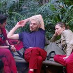Dschungelcamp-Szene: Alessia Herren (rechts) und Sam Dylan (Mitte) sitzen mit Yeliz Koc (links) auf einer Holzbank im Camp. Sie diskutieren intensiv, während Sam entspannt mit einer Hand hinter dem Kopf lehnt. Im Hintergrund ist der dichte Dschungel mit Pflanzen und einer Laterne zu sehen. Yeliz' Wasserflasche mit ihrem Namen steht neben ihr.
