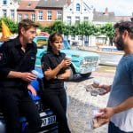"Friesland – Landfluchten": Die Polizisten Süher Özlügül (Sophie Dal) und Henk Cassens (Maxim Mehmet) lehnen an ihrem Streifenwagen. Yunus (Yunus Cumartpay) hat den beiden Kaffee und Brötchen mitgebracht und bietet sie ihnen an.