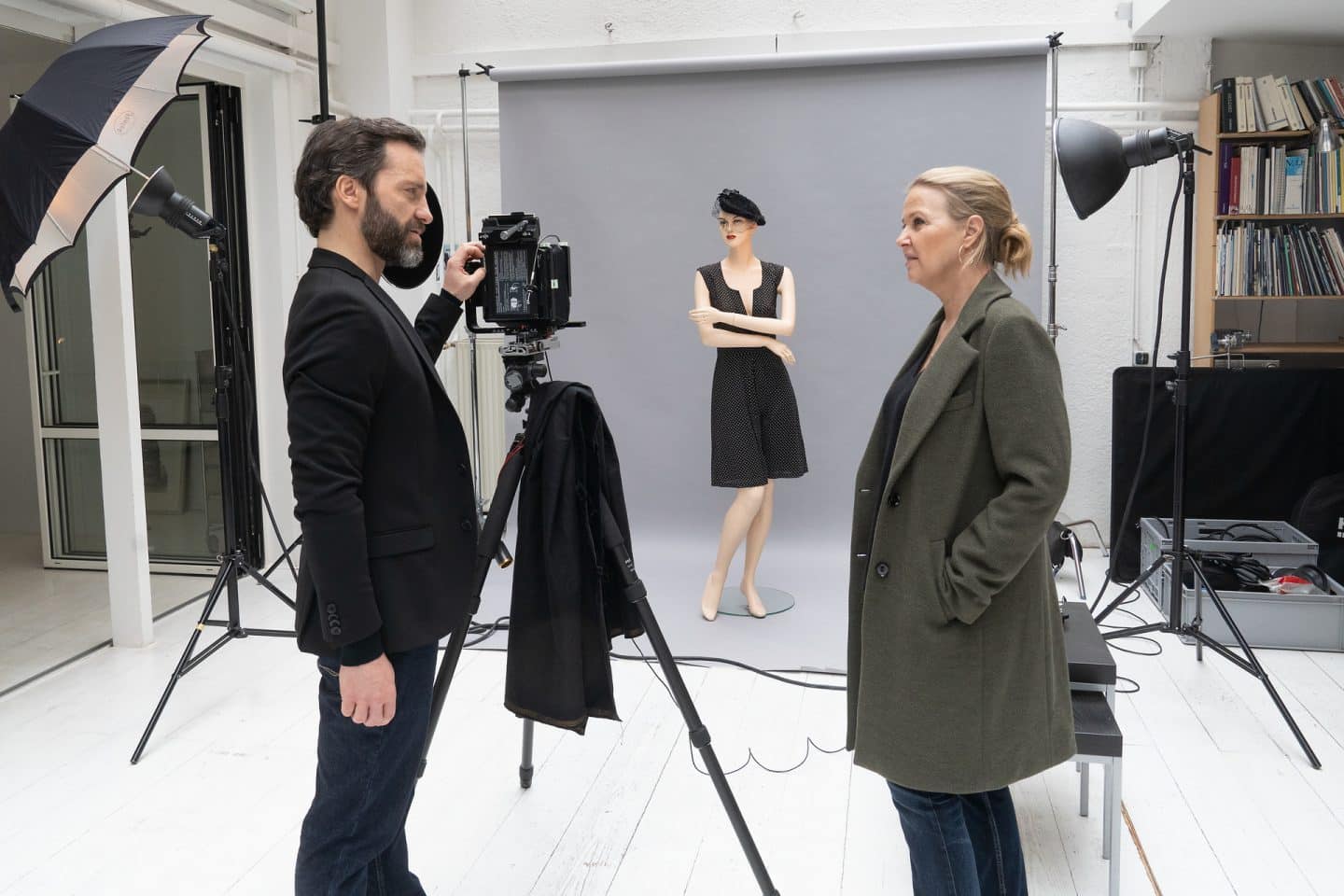 "Die Chefin - True Crime": In einem weißen Fotostudio befragt Vera Lanz (Katharina Böhm) den schwarz gekleideten Fotografen Ulf Brandt (Thomas Wodianka). Er hat eine Hand an seiner Kamera, die auf einem Stativ steht und auf eine ausgerollte graue Leinwand gerichtet ist. Dort steht eine Schaufensterpuppe mit verschränkten Armen, gekleidet in ein kurzes schwarzes Kleid und eine schwarze Baskenmütze.