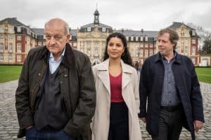 "Wilsberg - Über dem Gesetz": Wilsberg (Leonard Lansink), Dr. Tessa Tilker (Patricia Meeden) und Ekki Talkötter (Oliver Korittke) laufen über den Vorplatz eines Schlosses. Die Fassade des historischen Gebäudes erhebt sich im Hintergrund.