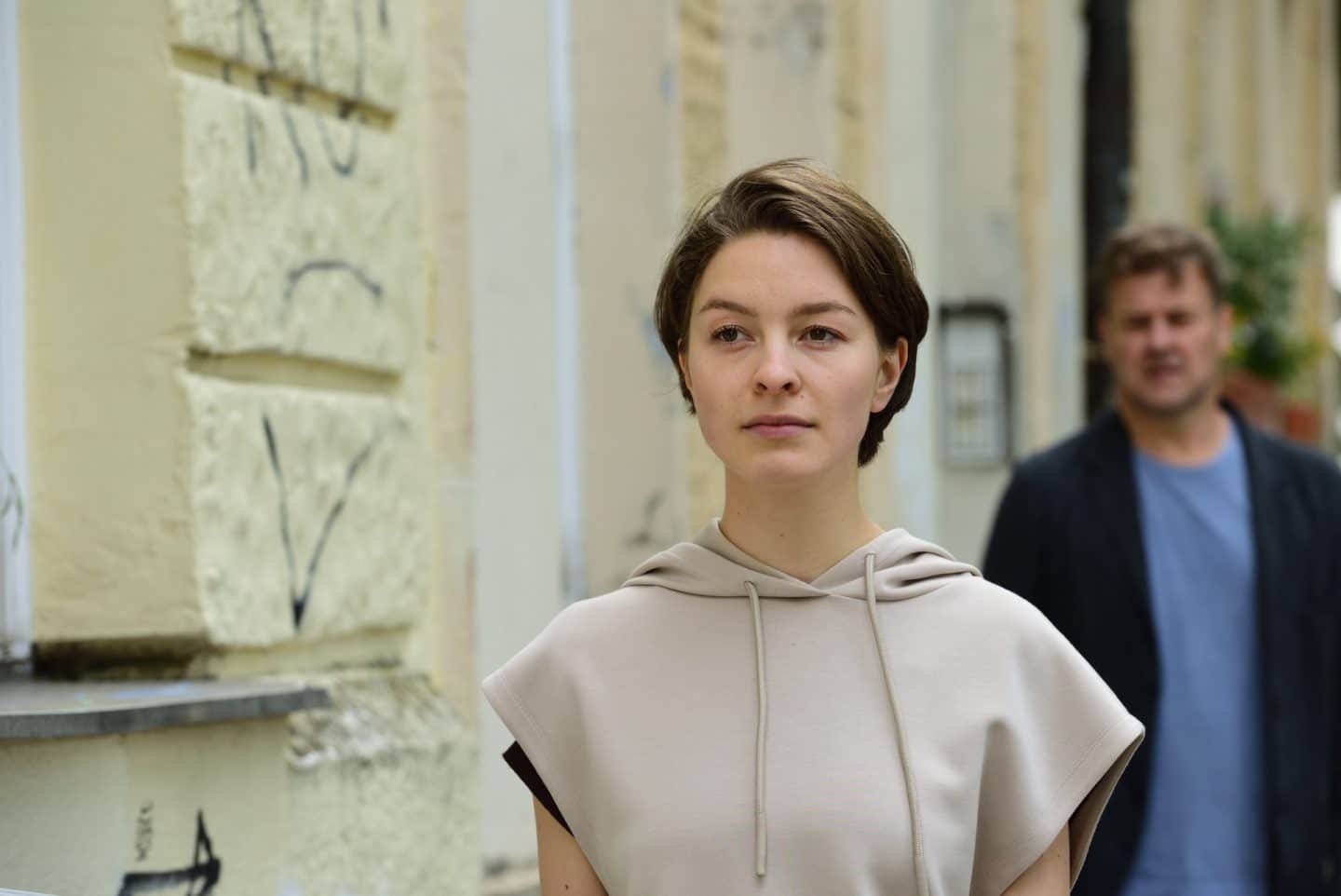 "München Mord - Nix für Angsthasen": Sara von Hohenburg (Maria Wördemann) läuft an einer Hausfassade entlang, während Harald (Marcus Mittermeier), der verschwommen im Hintergrund zu sehen ist, ihr nachschaut.

