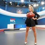 Moderatorin Jana Wosnitza steht im modernen NFL-Studio von RTL. Sie trägt einen schwarzen Blazer, schwarze Shorts und weiße Pumps und hält einen Football in beiden Händen vor sich. Im Hintergrund sind Bildschirme mit dem NFL-Logo und Grafiken zu sehen.