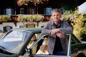 "Der Bergdoktor" - 18. Staffel: Vor der Arztpraxis: Dr. Martin Gruber (Hans Sigl) lehnt an der geöffneten Tür seines grünen Mercedes und lächelt in die Kamera.