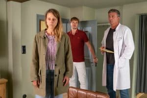 "Der Bergdoktor: Ein neuer Mensch": In der Arztpraxis: Blass, mit rot unterlaufenen Augen und hängenden Schultern steht Tina Reichert (Xenia Tiling) mit dem Rücken zu Dr. Martin Gruber (Hans Sigl). Dieser hält einen Umschlag in der Hand und blickt nachdenklich seine Patientin an. In der geöffneten Tür steht David Kästner (Frédéric Brossier) und wirkt irritiert.