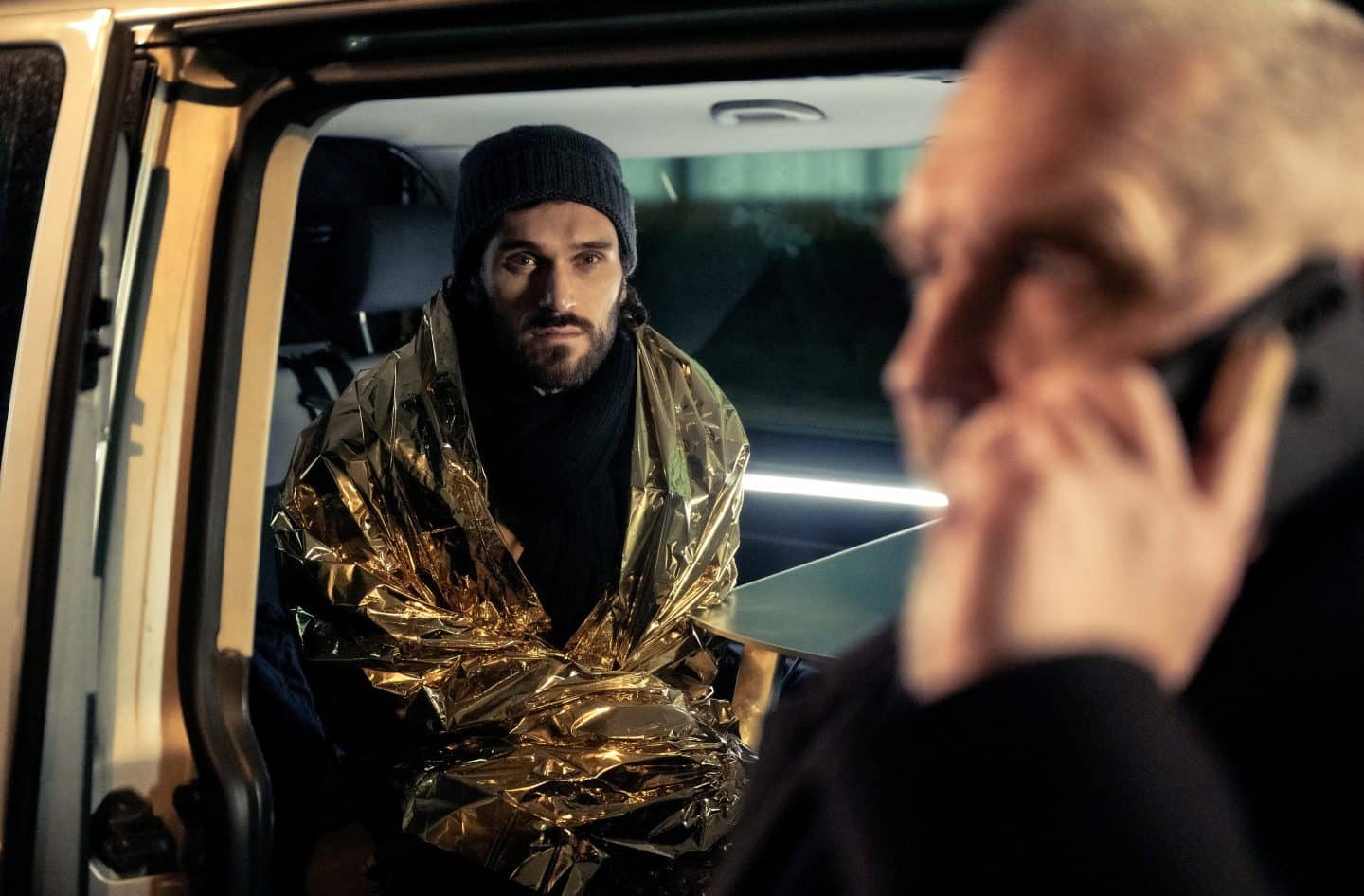 „Tatort: Restschuld“: Schuld, Blut und ein unsichtbares Opfer – Lohnt sich der neue Fall aus Köln? 1 Ein Mann mit dunklem Bart und Mütze (David Gross, gespielt von Vladimir Korneev) sitzt in einem Fahrzeug, in eine Rettungsdecke gehüllt, und blickt sichtlich geschockt in die Kamera. Im Vordergrund spricht eine verschwommene Person (Freddy Schenk, gespielt von Dietmar Bär) am Telefon. Die Szene zeigt eine Befragung am Tatort, bei der emotionaler Stress deutlich wird.