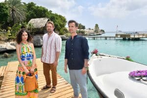 "Das Traumschiff: Curaçao": Am Meer: Neben einem Holzsteg liegt ein mit Blumen geschmücktes Boot. Auf dem Steg stehen Max Parger (Florian Silbereisen), Valerie Prinz (Nadine Menz) und Philipp Korte (David Korbmann). Traurig hält Valerie eine Tasche in den Händen.