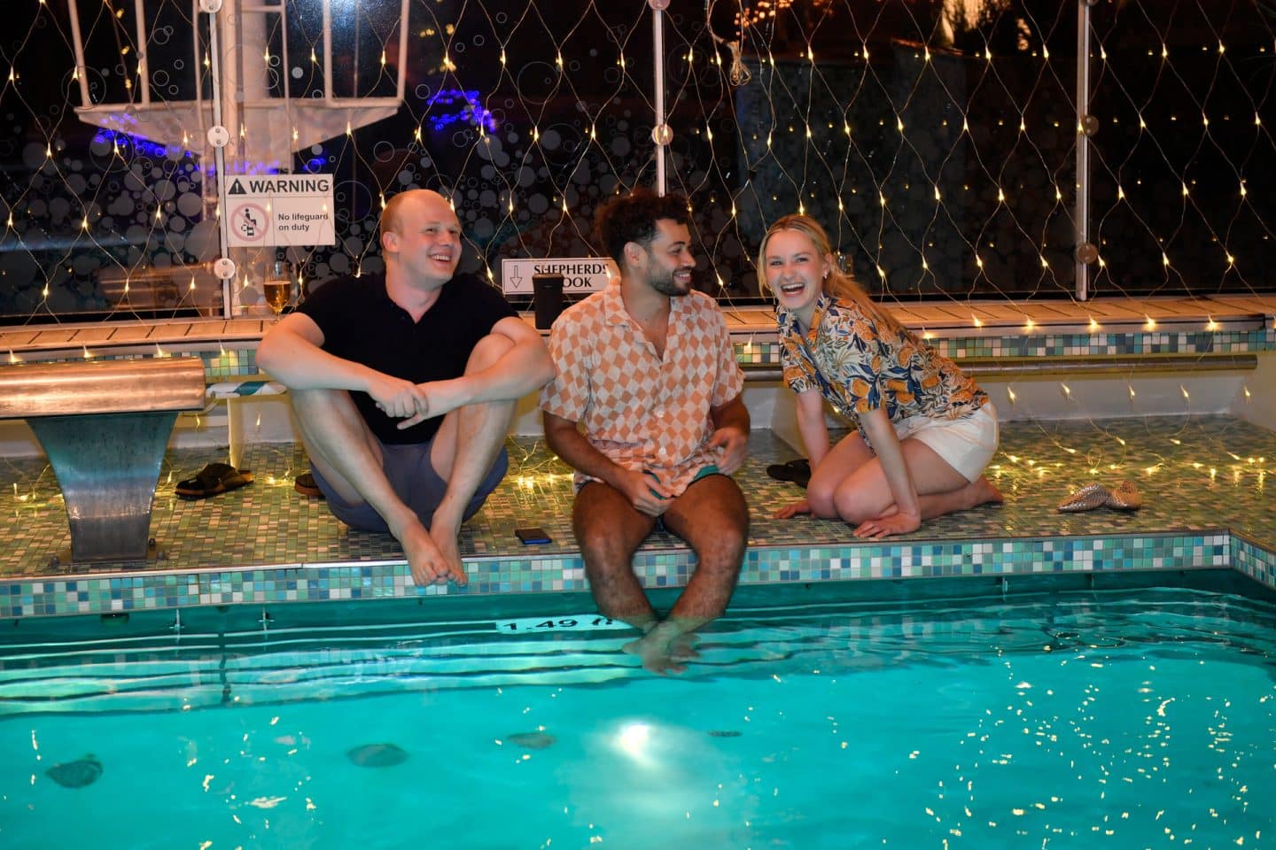 "Das Traumschiff: Curaçao": Am Abend: Florian Pfeiffer (Sebastian Jakob Doppelbauer), Malik Al Ghoul (Robin Cadet) und Liv Simonis (Pauline Werner) sitzen lachend am beleuchteten Schiffspool.
