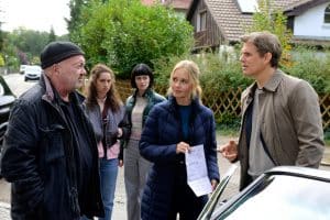 "Ein starkes Team - Der tote Mörder": Auf der Straße in einer Wohnsiedlung zeigt Linett (Stefanie Stappenbeck) Peter Perl (Steffen Groth) ein Schreiben der Staatsanwaltschaft. Beide sind im Gespräch und haben den Blick auf Otto (Florian Martens) gerichtet, der Peter zufrieden anlächelt. Im Hintergrund beobachten Karina (Joone Dankou) und Lotte (Sidney Fahlisch) die drei.