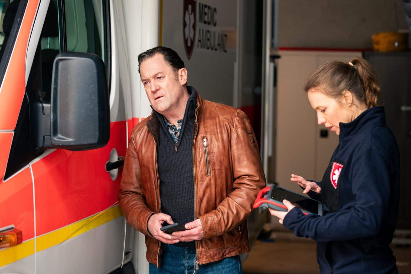 „Die Chefin – Grenzgänger“ – Mord, Drogen und ein dubioses Meditationszentrum 1 "Die Chefin - Grenzgänger": Paul Böhmer (Jürgen Tonkel) untersucht einen Schaden am Rückspiegel des Rettungswagens. Martina Jessing (Anna Brüggemann) steht neben ihm und prüft auf einem Tablet die Dokumentation.