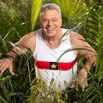 Jürgen Hingsen, ehemaliger deutscher Zehnkämpfer, in einem weißen Trikot mit rotem und schwarzem Streifen sowie gelbem Stern, lächelnd inmitten von grünem Dschungel.