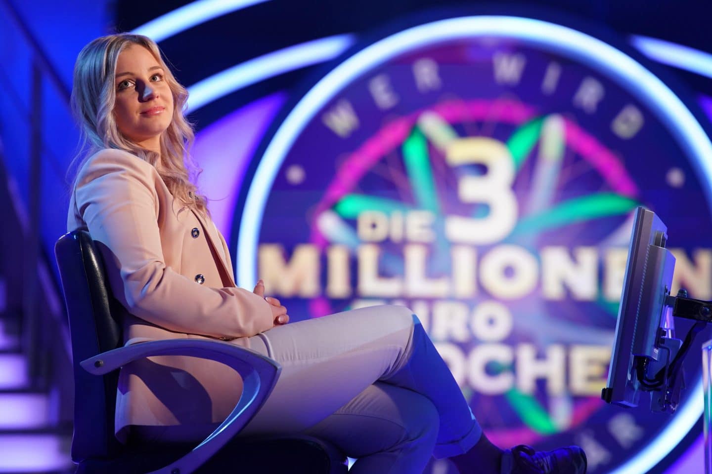 Laetitia Richter, Kandidatin bei 'Wer wird Millionär?', sitzt in einem eleganten rosa Blazer auf dem Stuhl der Showbühne vor dem Hintergrund der 3-Millionen-Euro-Woche, mit einem nachdenklichen und konzentrierten Blick. 
