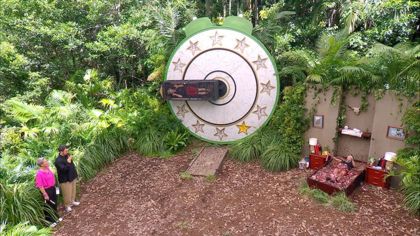 Dschungelcamp 2025: Edith Stehfest und Sam Dylan treten zur Dschungelprüfung 'Keine Uhrsache' an. Sam ist im rotierenden Zeiger einer riesigen Uhr gefangen, während Edith im Bett aus Fleischabfällen Fragen vorliest. Links beobachten die Moderatoren Sonja Zietlow und Jan Köppen das Geschehen.