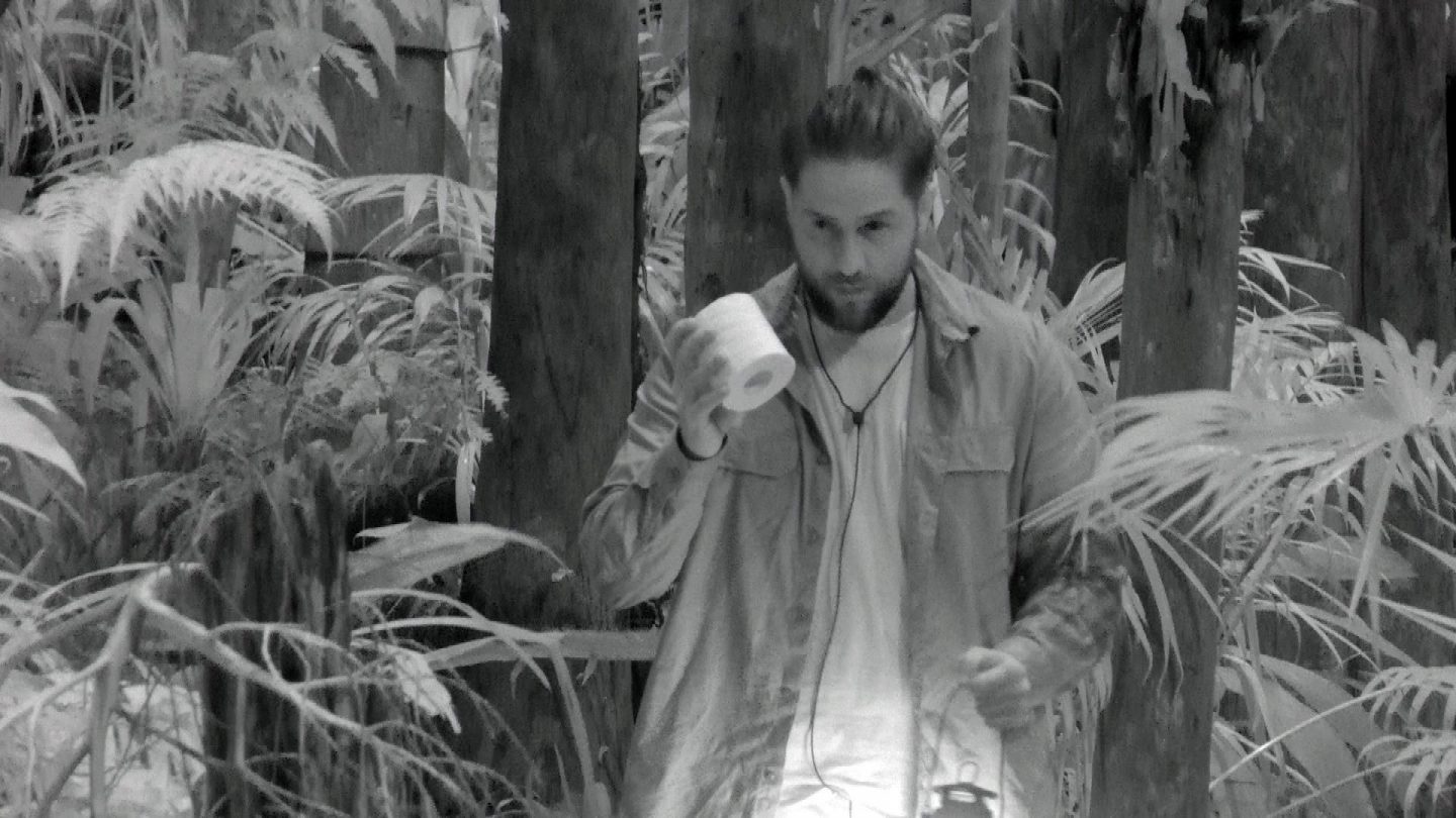 Maurice Dziwak im nächtlichen Dschungelcamp, aufgenommen in Schwarz-Weiß. Er hält eine Rolle Toilettenpapier in der Hand und blickt entschlossen in die Kamera. Maurice trägt ein helles Shirt und eine offene Jacke darüber, um den Hals hängt eine Kette. In der anderen Hand hält er eine Laterne, die seinen Weg durch die dichte Vegetation des Camps erleuchtet. Im Hintergrund sind Holzbalken und üppige Pflanzen zu sehen.