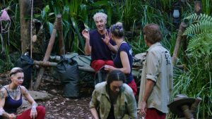 Sam Dylan sitzt auf einer Holzbank im Camp und hält ein abgebrochenes Stück seiner Make-up-Palette in der Hand. Er verteidigt sich gestikulierend gegenüber den anderen Camp-Bewohnern und beteuert, dass es sich um ein Versehen handelt. Skeptische Blicke zeigen jedoch, dass ihm nicht alle glauben. Die Diskussion um die Schmuggelware sorgt für Spannungen in der Gruppe.