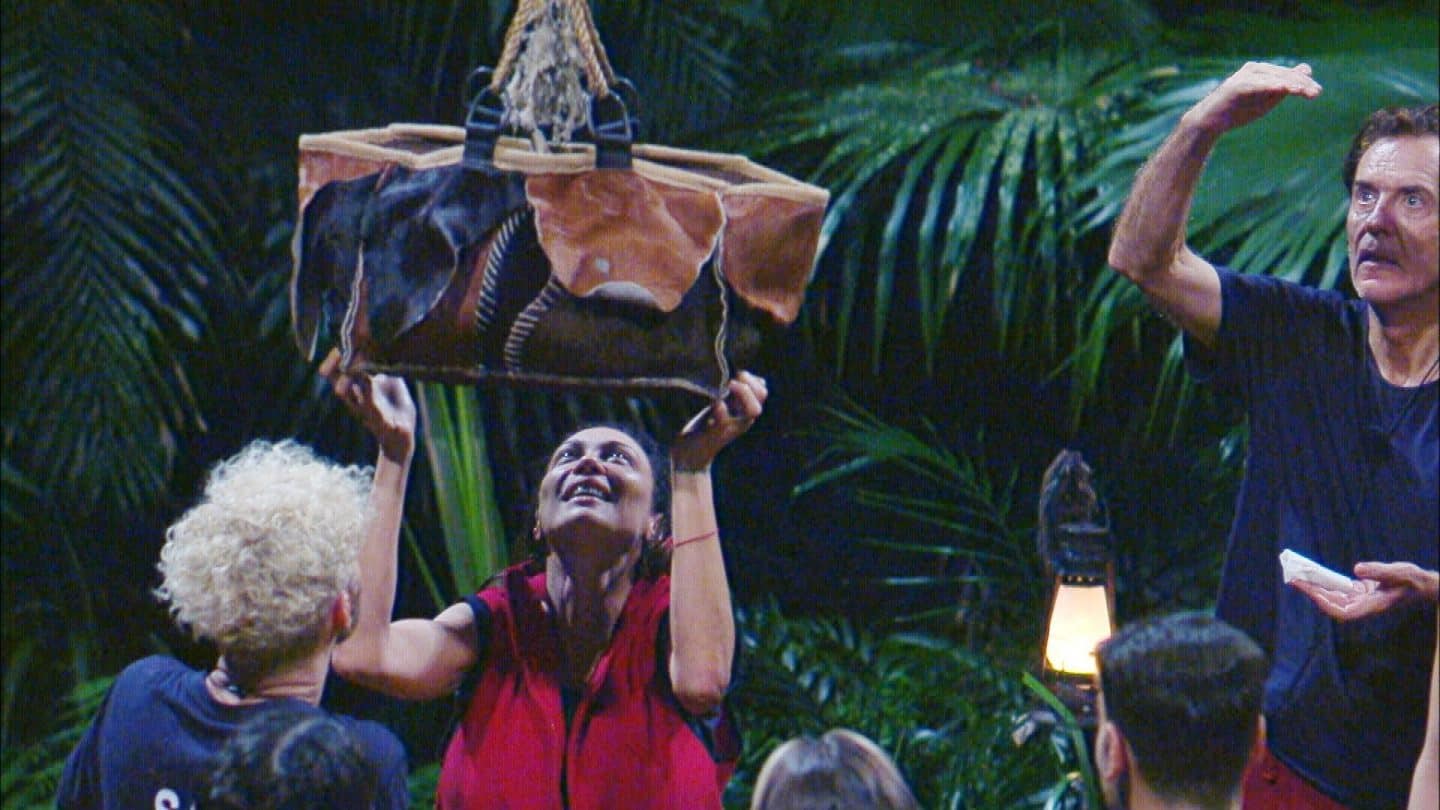 andidaten im Dschungelcamp erhalten eine Essenslieferung nach erfolgreicher Prüfung. Lilly Becker (Mitte) hebt einen geflochtenen Korb mit Lebensmitteln hoch, während Sam Dylan (links) und Jörg Dahlmann (rechts) das Geschehen gespannt beobachten. Der Dschungelhintergrund ist von dichter Vegetation umgeben, eine Laterne sorgt für stimmungsvolle Beleuchtung.