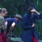 Anna-Carina Woitschack und Lilly Becker flüchten während eines starken Regenschauers im Dschungelcamp in das Dschungeltelefon. Anna-Carina trägt eine nasse beige Jacke mit ihrem Namen auf dem Rücken und eine rote Hose, während Lilly in einer dunklen Jacke Schutz sucht und ihre Hand über die Stirn hält, um sich vor dem prasselnden Regen zu schützen. Im Hintergrund ist dichter grüner Dschungel zu sehen.