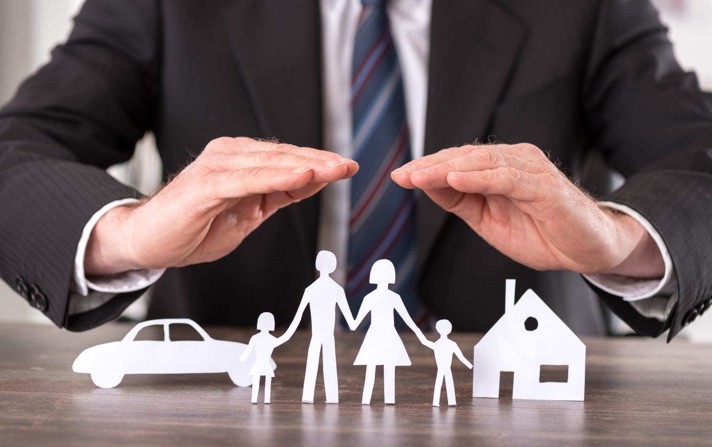 Ein Geschäftsmann in einem dunklen Anzug und Krawatte legt schützend beide Hände über symbolische Papierfiguren, die eine Familie mit zwei Kindern, ein Auto und ein Haus darstellen. Die Szene symbolisiert den Schutz und die Absicherung von Familie, Eigentum und Fahrzeug durch Versicherungen oder finanzielle Vorsorge.