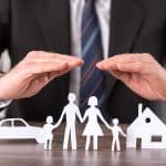 Ein Geschäftsmann in einem dunklen Anzug und Krawatte legt schützend beide Hände über symbolische Papierfiguren, die eine Familie mit zwei Kindern, ein Auto und ein Haus darstellen. Die Szene symbolisiert den Schutz und die Absicherung von Familie, Eigentum und Fahrzeug durch Versicherungen oder finanzielle Vorsorge.