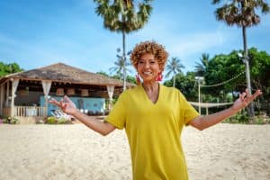 Arabella Kiesbauer steht in einem leuchtend gelben Kleid an einem sonnigen Strand vor einer strohgedeckten Hütte. Im Hintergrund sind Palmen und eine entspannte, tropische Atmosphäre zu sehen. Sie lächelt herzlich und breitet die Arme aus, was eine einladende und fröhliche Stimmung vermittelt.