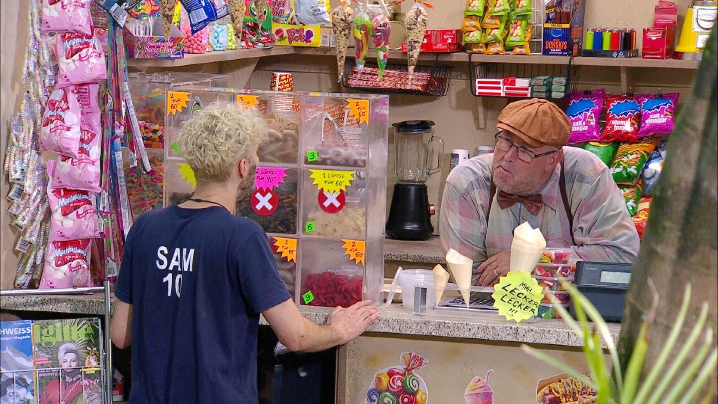 Sam Dylan steht im Dschungel-Kiosk vor dem Tresen und spricht mit Kioskbesitzer Matt, der hinter der Theke lehnt. Hinter ihm sind zahlreiche Süßigkeiten und Snacks aufgereiht, während in den Vorratsboxen der Ekelprüfung exotische Spezialitäten lagern. Sam wählt seine Herausforderung, während Matt ihn skeptisch mustert.