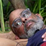 Pierre Sanoussi-Bliss liegt entspannt im Dschungelcamp auf einer Unterlage und lehnt seinen Kopf an einen Baumstamm. Mit nachdenklichem Blick und durchsichtiger Brille scheint er in die Ferne zu schauen.