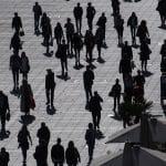 Menschenmenge auf einem sonnigen Platz: Silhouetten von Personen, die in unterschiedliche Richtungen gehen, mit Schatten auf dem Boden und einem urbanen Hintergrund