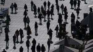 Menschenmenge auf einem sonnigen Platz: Silhouetten von Personen, die in unterschiedliche Richtungen gehen, mit Schatten auf dem Boden und einem urbanen Hintergrund