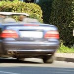 Ein dunkelblaues Cabrio fährt mit hoher Geschwindigkeit an einer versteckten mobilen Radarkamera vorbei, die am Straßenrand hinter einer Hecke positioniert ist.
