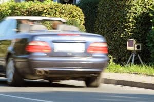 Ein dunkelblaues Cabrio fährt mit hoher Geschwindigkeit an einer versteckten mobilen Radarkamera vorbei, die am Straßenrand hinter einer Hecke positioniert ist.