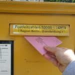 Ein gelber Briefkasten der Deutschen Post mit einem Fach für Postleitzahlen der Region Berlin/Brandenburg. Eine Hand schiebt einen rosafarbenen Brief mit der Aufschrift "Wahlbrief" in den Schlitz. Im unteren Bereich sind die Leerungszeiten des Briefkastens auf einem Schild zu erkennen. Im Hintergrund ist unscharf eine grüne Parkanlage zu sehen.
