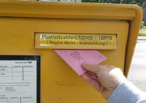 Ein gelber Briefkasten der Deutschen Post mit einem Fach für Postleitzahlen der Region Berlin/Brandenburg. Eine Hand schiebt einen rosafarbenen Brief mit der Aufschrift "Wahlbrief" in den Schlitz. Im unteren Bereich sind die Leerungszeiten des Briefkastens auf einem Schild zu erkennen. Im Hintergrund ist unscharf eine grüne Parkanlage zu sehen.