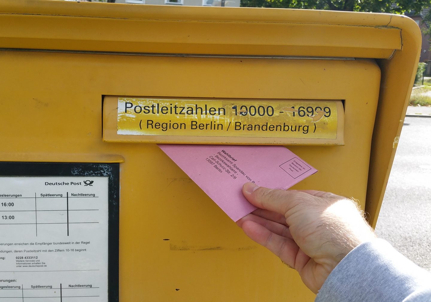 Ein gelber Briefkasten der Deutschen Post mit einem Fach für Postleitzahlen der Region Berlin/Brandenburg. Eine Hand schiebt einen rosafarbenen Brief mit der Aufschrift "Wahlbrief" in den Schlitz. Im unteren Bereich sind die Leerungszeiten des Briefkastens auf einem Schild zu erkennen. Im Hintergrund ist unscharf eine grüne Parkanlage zu sehen.