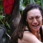 Lilly Becker sitzt lachend im Dschungelcamp vor tropischer Kulisse. Sie trägt ein schwarzes Oberteil und hat ihre Haare in Zöpfen geflochten. Im Hintergrund sind ein Rucksack und grüne Blätter zu sehen.