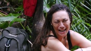 Lilly Becker sitzt lachend im Dschungelcamp vor tropischer Kulisse. Sie trägt ein schwarzes Oberteil und hat ihre Haare in Zöpfen geflochten. Im Hintergrund sind ein Rucksack und grüne Blätter zu sehen.