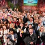 Gruppenfoto mit Teilnehmern aus 20 Jahren „Bauer sucht Frau“. Männer und Frauen stoßen fröhlich mit Gläsern an, um das Jubiläum der beliebten TV-Sendung zu feiern. Im Hintergrund ist eine festliche Kulisse mit Heuballen, einem roten Teppich und einer Leuchtschrift „LOVE“ zu sehen.