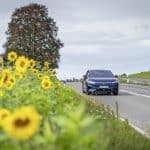 Der vollelektrische VW ID.7 Pro S fährt auf einer idyllischen Landstraße entlang eines blühenden Sonnenblumenfelds. Im Hintergrund sind ein traditionelles Landhaus und ein großer Baum zu sehen, während der Himmel von leichten Wolken bedeckt ist.