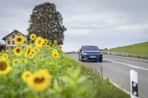 Der vollelektrische VW ID.7 Pro S fährt auf einer idyllischen Landstraße entlang eines blühenden Sonnenblumenfelds. Im Hintergrund sind ein traditionelles Landhaus und ein großer Baum zu sehen, während der Himmel von leichten Wolken bedeckt ist.