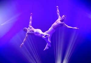 Devin & Davide präsentieren eine atemberaubende Luftakrobatik-Show an Strapaten. Beide Künstler, in weißen Kostümen, führen synchron dynamische Bewegungen in schwindelerregender Höhe aus, umgeben von intensivem lila Bühnenlicht. Strahlen im Hintergrund verstärken die Dramatik und Eleganz ihrer Darbietung.