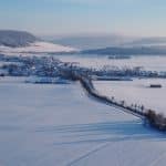 Verschneite Landschaft mit einer kleinen, malerischen Ortschaft, umgeben von weitläufigen Feldern und sanften Hügeln unter klarem blauem Himmel.