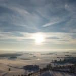 "Winterliche Landschaft mit schneebedeckten Feldern und einer Ortschaft im Hintergrund, unter klarem Himmel und Sonnenschein."