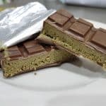 Eine aufgebrochene Tafel Schokolade mit einer grünen, krümeligen Füllung liegt auf einem weißen Teller. Die silberne Verpackung ist teilweise geöffnet, und die Schokolade hat eine glatte, glänzende Oberfläche mit quadratischem Muster. Die Füllung könnte aus Matcha oder Pistazien bestehen.