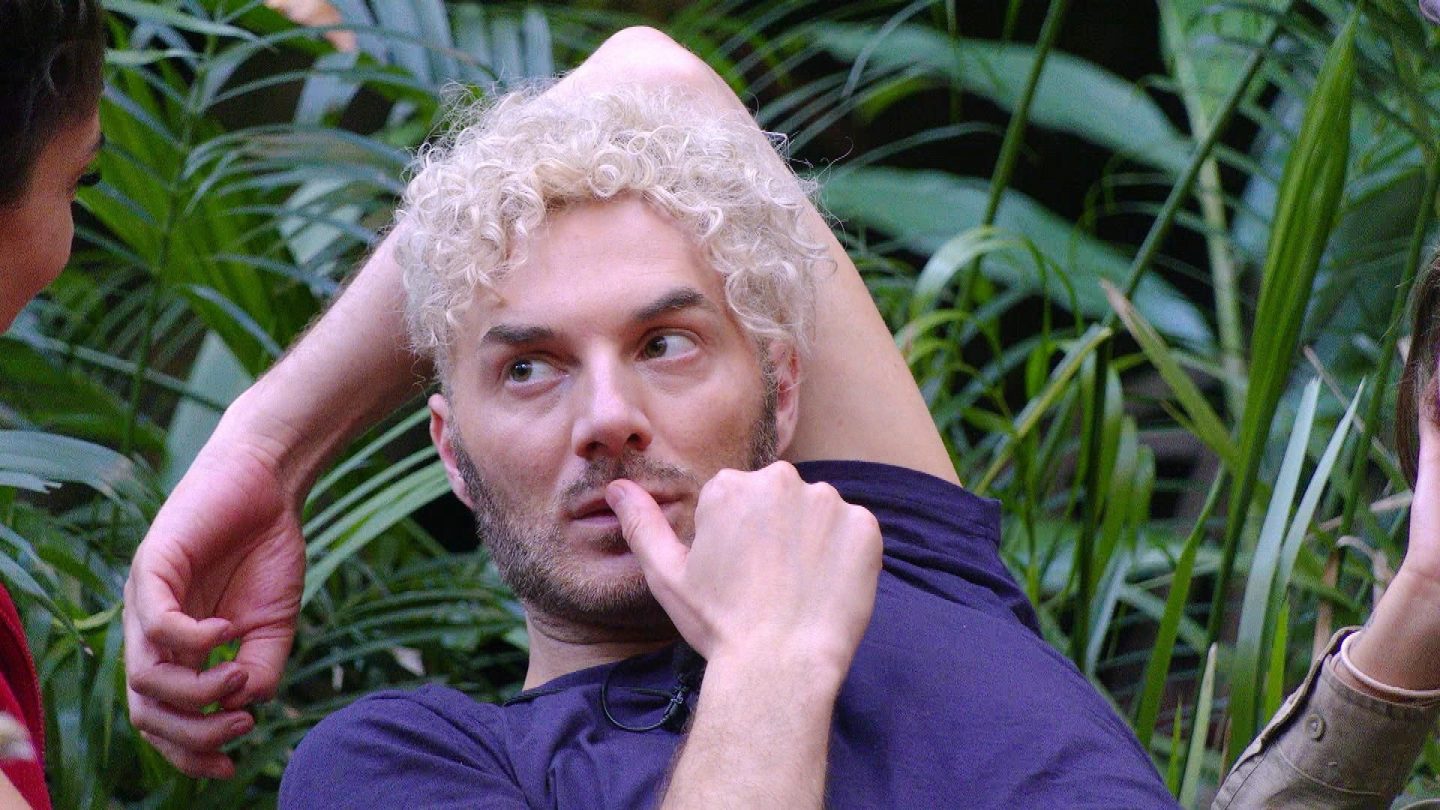 Nahaufnahme von Sam Dylan im Dschungelcamp. Mit nachdenklichem Blick und Hand am Mund hört er aufmerksam zu, während Alessia Herren und Yeliz Koc mit ihm über den aktuellen Stand zwischen Yeliz und Jimi Blue Ochsenknecht sprechen. Im Hintergrund ist dichte grüne Vegetation zu sehen.