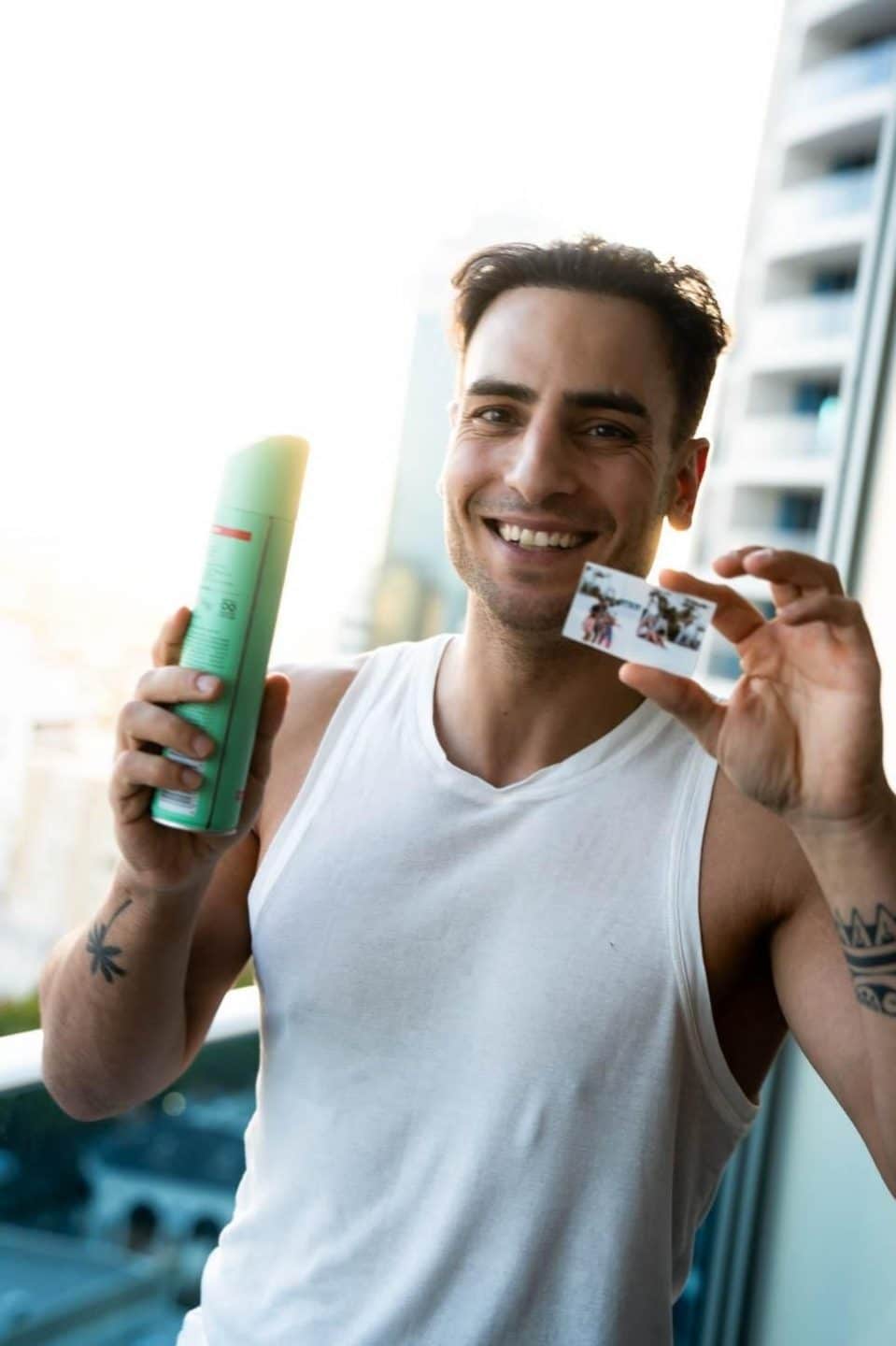 Timur Ülker hält eine Dose Haarspray und ein kleines Familienfoto in den Händen und lächelt in die Kamera.

